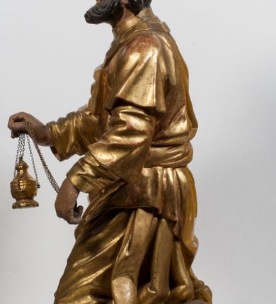 Zdjęcie nr 25: Tabernakulum w formie Arki Przymierza, adorowanej przez dwie klęczące figury starotestamentowego arcykapłana i biskupa oraz dwie stojące postacie lewitów oraz klęczące aniołów, zwieńczonej glorią promienistą z okiem Opatrzności. Wsparte na ośmiu cylindrycznych nóżkach prostopadłościenne tabernakulum jest od frontu podzielone na trzy pola wyznaczone kanelowanymi listwami. Pole środkowe stanowią drzwiczki z rytymi cyframi Dekalogu z ukrzyżowanym Chrystusem, pola boczne z wiązką pałek wodnych z jednej i winnym gronem z drugiej strony. Na skłębionych chmurach z podestem leży Baranek (obecnie w jego miejscu znajduje się figura Matki Boskiej). W tle znacznej wielkości gloria promienista z wieńcem chmur i umieszczonym w nim okiem Opatrzności oraz dwie uskrzydlone główki, nad którymi papieska tiara ze skrzyżowanymi kluczami. Po bokach Baranka dwa klęczące, uskrzydlone nagie aniołki. Obok tabernakulum dwie klęczące postacie z kadzielnicami w rękach. Postać po lewej w stroju biskupim, w albie, stule i kapie oraz infule z dwiema taśmami. Postać po prawej stronie jest ubrany w tradycyjny starotestamentowy strój – efod czyli tunikę i płaszcz wierzchni przepasany na biodrach, pektorał na piersi i zawój na głowie. Za nimi dwaj stojący lewici w tunikach przepasanych na biodrach i wysokich butach. Adorujące Baranka anioły w pozycji klęczącej z rękami skrzyżowanymi na piersi, ze skrzydłami na plecach i nogami w połowie utworzonymi ze skrzydeł.  


