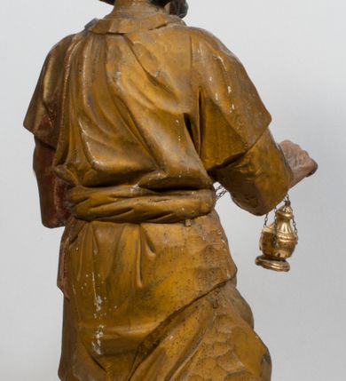 Zdjęcie nr 29: Tabernakulum w formie Arki Przymierza, adorowanej przez dwie klęczące figury starotestamentowego arcykapłana i biskupa oraz dwie stojące postacie lewitów oraz klęczące aniołów, zwieńczonej glorią promienistą z okiem Opatrzności. Wsparte na ośmiu cylindrycznych nóżkach prostopadłościenne tabernakulum jest od frontu podzielone na trzy pola wyznaczone kanelowanymi listwami. Pole środkowe stanowią drzwiczki z rytymi cyframi Dekalogu z ukrzyżowanym Chrystusem, pola boczne z wiązką pałek wodnych z jednej i winnym gronem z drugiej strony. Na skłębionych chmurach z podestem leży Baranek (obecnie w jego miejscu znajduje się figura Matki Boskiej). W tle znacznej wielkości gloria promienista z wieńcem chmur i umieszczonym w nim okiem Opatrzności oraz dwie uskrzydlone główki, nad którymi papieska tiara ze skrzyżowanymi kluczami. Po bokach Baranka dwa klęczące, uskrzydlone nagie aniołki. Obok tabernakulum dwie klęczące postacie z kadzielnicami w rękach. Postać po lewej w stroju biskupim, w albie, stule i kapie oraz infule z dwiema taśmami. Postać po prawej stronie jest ubrany w tradycyjny starotestamentowy strój – efod czyli tunikę i płaszcz wierzchni przepasany na biodrach, pektorał na piersi i zawój na głowie. Za nimi dwaj stojący lewici w tunikach przepasanych na biodrach i wysokich butach. Adorujące Baranka anioły w pozycji klęczącej z rękami skrzyżowanymi na piersi, ze skrzydłami na plecach i nogami w połowie utworzonymi ze skrzydeł.  


