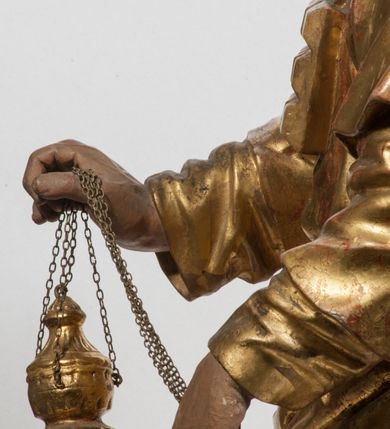 Zdjęcie nr 23: Tabernakulum w formie Arki Przymierza, adorowanej przez dwie klęczące figury starotestamentowego arcykapłana i biskupa oraz dwie stojące postacie lewitów oraz klęczące aniołów, zwieńczonej glorią promienistą z okiem Opatrzności. Wsparte na ośmiu cylindrycznych nóżkach prostopadłościenne tabernakulum jest od frontu podzielone na trzy pola wyznaczone kanelowanymi listwami. Pole środkowe stanowią drzwiczki z rytymi cyframi Dekalogu z ukrzyżowanym Chrystusem, pola boczne z wiązką pałek wodnych z jednej i winnym gronem z drugiej strony. Na skłębionych chmurach z podestem leży Baranek (obecnie w jego miejscu znajduje się figura Matki Boskiej). W tle znacznej wielkości gloria promienista z wieńcem chmur i umieszczonym w nim okiem Opatrzności oraz dwie uskrzydlone główki, nad którymi papieska tiara ze skrzyżowanymi kluczami. Po bokach Baranka dwa klęczące, uskrzydlone nagie aniołki. Obok tabernakulum dwie klęczące postacie z kadzielnicami w rękach. Postać po lewej w stroju biskupim, w albie, stule i kapie oraz infule z dwiema taśmami. Postać po prawej stronie jest ubrany w tradycyjny starotestamentowy strój – efod czyli tunikę i płaszcz wierzchni przepasany na biodrach, pektorał na piersi i zawój na głowie. Za nimi dwaj stojący lewici w tunikach przepasanych na biodrach i wysokich butach. Adorujące Baranka anioły w pozycji klęczącej z rękami skrzyżowanymi na piersi, ze skrzydłami na plecach i nogami w połowie utworzonymi ze skrzydeł.  


