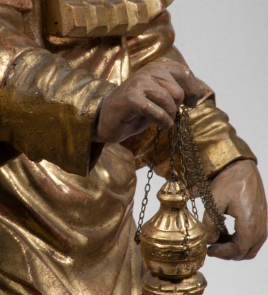 Zdjęcie nr 32: Tabernakulum w formie Arki Przymierza, adorowanej przez dwie klęczące figury starotestamentowego arcykapłana i biskupa oraz dwie stojące postacie lewitów oraz klęczące aniołów, zwieńczonej glorią promienistą z okiem Opatrzności. Wsparte na ośmiu cylindrycznych nóżkach prostopadłościenne tabernakulum jest od frontu podzielone na trzy pola wyznaczone kanelowanymi listwami. Pole środkowe stanowią drzwiczki z rytymi cyframi Dekalogu z ukrzyżowanym Chrystusem, pola boczne z wiązką pałek wodnych z jednej i winnym gronem z drugiej strony. Na skłębionych chmurach z podestem leży Baranek (obecnie w jego miejscu znajduje się figura Matki Boskiej). W tle znacznej wielkości gloria promienista z wieńcem chmur i umieszczonym w nim okiem Opatrzności oraz dwie uskrzydlone główki, nad którymi papieska tiara ze skrzyżowanymi kluczami. Po bokach Baranka dwa klęczące, uskrzydlone nagie aniołki. Obok tabernakulum dwie klęczące postacie z kadzielnicami w rękach. Postać po lewej w stroju biskupim, w albie, stule i kapie oraz infule z dwiema taśmami. Postać po prawej stronie jest ubrany w tradycyjny starotestamentowy strój – efod czyli tunikę i płaszcz wierzchni przepasany na biodrach, pektorał na piersi i zawój na głowie. Za nimi dwaj stojący lewici w tunikach przepasanych na biodrach i wysokich butach. Adorujące Baranka anioły w pozycji klęczącej z rękami skrzyżowanymi na piersi, ze skrzydłami na plecach i nogami w połowie utworzonymi ze skrzydeł.  


