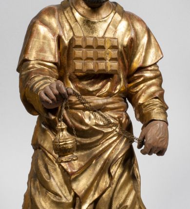 Zdjęcie nr 38: Tabernakulum w formie Arki Przymierza, adorowanej przez dwie klęczące figury starotestamentowego arcykapłana i biskupa oraz dwie stojące postacie lewitów oraz klęczące aniołów, zwieńczonej glorią promienistą z okiem Opatrzności. Wsparte na ośmiu cylindrycznych nóżkach prostopadłościenne tabernakulum jest od frontu podzielone na trzy pola wyznaczone kanelowanymi listwami. Pole środkowe stanowią drzwiczki z rytymi cyframi Dekalogu z ukrzyżowanym Chrystusem, pola boczne z wiązką pałek wodnych z jednej i winnym gronem z drugiej strony. Na skłębionych chmurach z podestem leży Baranek (obecnie w jego miejscu znajduje się figura Matki Boskiej). W tle znacznej wielkości gloria promienista z wieńcem chmur i umieszczonym w nim okiem Opatrzności oraz dwie uskrzydlone główki, nad którymi papieska tiara ze skrzyżowanymi kluczami. Po bokach Baranka dwa klęczące, uskrzydlone nagie aniołki. Obok tabernakulum dwie klęczące postacie z kadzielnicami w rękach. Postać po lewej w stroju biskupim, w albie, stule i kapie oraz infule z dwiema taśmami. Postać po prawej stronie jest ubrany w tradycyjny starotestamentowy strój – efod czyli tunikę i płaszcz wierzchni przepasany na biodrach, pektorał na piersi i zawój na głowie. Za nimi dwaj stojący lewici w tunikach przepasanych na biodrach i wysokich butach. Adorujące Baranka anioły w pozycji klęczącej z rękami skrzyżowanymi na piersi, ze skrzydłami na plecach i nogami w połowie utworzonymi ze skrzydeł.  


