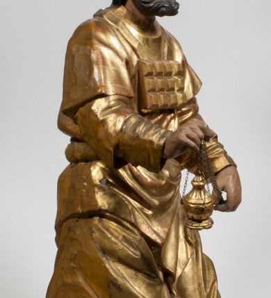 Zdjęcie nr 35: Tabernakulum w formie Arki Przymierza, adorowanej przez dwie klęczące figury starotestamentowego arcykapłana i biskupa oraz dwie stojące postacie lewitów oraz klęczące aniołów, zwieńczonej glorią promienistą z okiem Opatrzności. Wsparte na ośmiu cylindrycznych nóżkach prostopadłościenne tabernakulum jest od frontu podzielone na trzy pola wyznaczone kanelowanymi listwami. Pole środkowe stanowią drzwiczki z rytymi cyframi Dekalogu z ukrzyżowanym Chrystusem, pola boczne z wiązką pałek wodnych z jednej i winnym gronem z drugiej strony. Na skłębionych chmurach z podestem leży Baranek (obecnie w jego miejscu znajduje się figura Matki Boskiej). W tle znacznej wielkości gloria promienista z wieńcem chmur i umieszczonym w nim okiem Opatrzności oraz dwie uskrzydlone główki, nad którymi papieska tiara ze skrzyżowanymi kluczami. Po bokach Baranka dwa klęczące, uskrzydlone nagie aniołki. Obok tabernakulum dwie klęczące postacie z kadzielnicami w rękach. Postać po lewej w stroju biskupim, w albie, stule i kapie oraz infule z dwiema taśmami. Postać po prawej stronie jest ubrany w tradycyjny starotestamentowy strój – efod czyli tunikę i płaszcz wierzchni przepasany na biodrach, pektorał na piersi i zawój na głowie. Za nimi dwaj stojący lewici w tunikach przepasanych na biodrach i wysokich butach. Adorujące Baranka anioły w pozycji klęczącej z rękami skrzyżowanymi na piersi, ze skrzydłami na plecach i nogami w połowie utworzonymi ze skrzydeł.  


