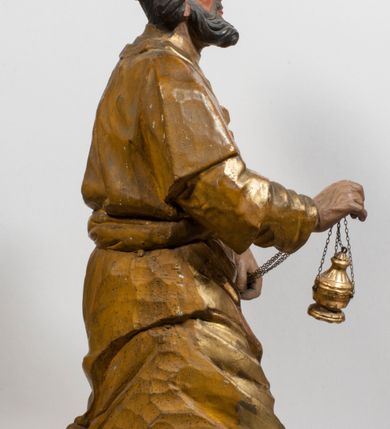 Zdjęcie nr 34: Tabernakulum w formie Arki Przymierza, adorowanej przez dwie klęczące figury starotestamentowego arcykapłana i biskupa oraz dwie stojące postacie lewitów oraz klęczące aniołów, zwieńczonej glorią promienistą z okiem Opatrzności. Wsparte na ośmiu cylindrycznych nóżkach prostopadłościenne tabernakulum jest od frontu podzielone na trzy pola wyznaczone kanelowanymi listwami. Pole środkowe stanowią drzwiczki z rytymi cyframi Dekalogu z ukrzyżowanym Chrystusem, pola boczne z wiązką pałek wodnych z jednej i winnym gronem z drugiej strony. Na skłębionych chmurach z podestem leży Baranek (obecnie w jego miejscu znajduje się figura Matki Boskiej). W tle znacznej wielkości gloria promienista z wieńcem chmur i umieszczonym w nim okiem Opatrzności oraz dwie uskrzydlone główki, nad którymi papieska tiara ze skrzyżowanymi kluczami. Po bokach Baranka dwa klęczące, uskrzydlone nagie aniołki. Obok tabernakulum dwie klęczące postacie z kadzielnicami w rękach. Postać po lewej w stroju biskupim, w albie, stule i kapie oraz infule z dwiema taśmami. Postać po prawej stronie jest ubrany w tradycyjny starotestamentowy strój – efod czyli tunikę i płaszcz wierzchni przepasany na biodrach, pektorał na piersi i zawój na głowie. Za nimi dwaj stojący lewici w tunikach przepasanych na biodrach i wysokich butach. Adorujące Baranka anioły w pozycji klęczącej z rękami skrzyżowanymi na piersi, ze skrzydłami na plecach i nogami w połowie utworzonymi ze skrzydeł.  


