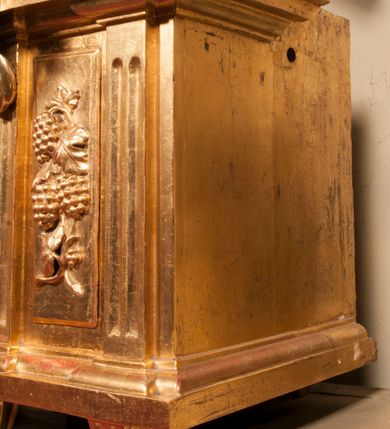 Zdjęcie nr 67: Tabernakulum w formie Arki Przymierza, adorowanej przez dwie klęczące figury starotestamentowego arcykapłana i biskupa oraz dwie stojące postacie lewitów oraz klęczące aniołów, zwieńczonej glorią promienistą z okiem Opatrzności. Wsparte na ośmiu cylindrycznych nóżkach prostopadłościenne tabernakulum jest od frontu podzielone na trzy pola wyznaczone kanelowanymi listwami. Pole środkowe stanowią drzwiczki z rytymi cyframi Dekalogu z ukrzyżowanym Chrystusem, pola boczne z wiązką pałek wodnych z jednej i winnym gronem z drugiej strony. Na skłębionych chmurach z podestem leży Baranek (obecnie w jego miejscu znajduje się figura Matki Boskiej). W tle znacznej wielkości gloria promienista z wieńcem chmur i umieszczonym w nim okiem Opatrzności oraz dwie uskrzydlone główki, nad którymi papieska tiara ze skrzyżowanymi kluczami. Po bokach Baranka dwa klęczące, uskrzydlone nagie aniołki. Obok tabernakulum dwie klęczące postacie z kadzielnicami w rękach. Postać po lewej w stroju biskupim, w albie, stule i kapie oraz infule z dwiema taśmami. Postać po prawej stronie jest ubrany w tradycyjny starotestamentowy strój – efod czyli tunikę i płaszcz wierzchni przepasany na biodrach, pektorał na piersi i zawój na głowie. Za nimi dwaj stojący lewici w tunikach przepasanych na biodrach i wysokich butach. Adorujące Baranka anioły w pozycji klęczącej z rękami skrzyżowanymi na piersi, ze skrzydłami na plecach i nogami w połowie utworzonymi ze skrzydeł.  


