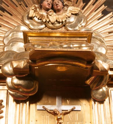 Zdjęcie nr 71: Tabernakulum w formie Arki Przymierza, adorowanej przez dwie klęczące figury starotestamentowego arcykapłana i biskupa oraz dwie stojące postacie lewitów oraz klęczące aniołów, zwieńczonej glorią promienistą z okiem Opatrzności. Wsparte na ośmiu cylindrycznych nóżkach prostopadłościenne tabernakulum jest od frontu podzielone na trzy pola wyznaczone kanelowanymi listwami. Pole środkowe stanowią drzwiczki z rytymi cyframi Dekalogu z ukrzyżowanym Chrystusem, pola boczne z wiązką pałek wodnych z jednej i winnym gronem z drugiej strony. Na skłębionych chmurach z podestem leży Baranek (obecnie w jego miejscu znajduje się figura Matki Boskiej). W tle znacznej wielkości gloria promienista z wieńcem chmur i umieszczonym w nim okiem Opatrzności oraz dwie uskrzydlone główki, nad którymi papieska tiara ze skrzyżowanymi kluczami. Po bokach Baranka dwa klęczące, uskrzydlone nagie aniołki. Obok tabernakulum dwie klęczące postacie z kadzielnicami w rękach. Postać po lewej w stroju biskupim, w albie, stule i kapie oraz infule z dwiema taśmami. Postać po prawej stronie jest ubrany w tradycyjny starotestamentowy strój – efod czyli tunikę i płaszcz wierzchni przepasany na biodrach, pektorał na piersi i zawój na głowie. Za nimi dwaj stojący lewici w tunikach przepasanych na biodrach i wysokich butach. Adorujące Baranka anioły w pozycji klęczącej z rękami skrzyżowanymi na piersi, ze skrzydłami na plecach i nogami w połowie utworzonymi ze skrzydeł.  


