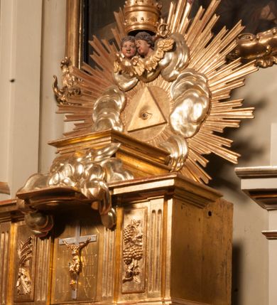 Zdjęcie nr 69: Tabernakulum w formie Arki Przymierza, adorowanej przez dwie klęczące figury starotestamentowego arcykapłana i biskupa oraz dwie stojące postacie lewitów oraz klęczące aniołów, zwieńczonej glorią promienistą z okiem Opatrzności. Wsparte na ośmiu cylindrycznych nóżkach prostopadłościenne tabernakulum jest od frontu podzielone na trzy pola wyznaczone kanelowanymi listwami. Pole środkowe stanowią drzwiczki z rytymi cyframi Dekalogu z ukrzyżowanym Chrystusem, pola boczne z wiązką pałek wodnych z jednej i winnym gronem z drugiej strony. Na skłębionych chmurach z podestem leży Baranek (obecnie w jego miejscu znajduje się figura Matki Boskiej). W tle znacznej wielkości gloria promienista z wieńcem chmur i umieszczonym w nim okiem Opatrzności oraz dwie uskrzydlone główki, nad którymi papieska tiara ze skrzyżowanymi kluczami. Po bokach Baranka dwa klęczące, uskrzydlone nagie aniołki. Obok tabernakulum dwie klęczące postacie z kadzielnicami w rękach. Postać po lewej w stroju biskupim, w albie, stule i kapie oraz infule z dwiema taśmami. Postać po prawej stronie jest ubrany w tradycyjny starotestamentowy strój – efod czyli tunikę i płaszcz wierzchni przepasany na biodrach, pektorał na piersi i zawój na głowie. Za nimi dwaj stojący lewici w tunikach przepasanych na biodrach i wysokich butach. Adorujące Baranka anioły w pozycji klęczącej z rękami skrzyżowanymi na piersi, ze skrzydłami na plecach i nogami w połowie utworzonymi ze skrzydeł.  



