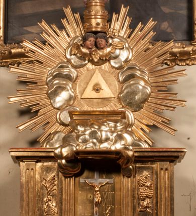 Zdjęcie nr 72: Tabernakulum w formie Arki Przymierza, adorowanej przez dwie klęczące figury starotestamentowego arcykapłana i biskupa oraz dwie stojące postacie lewitów oraz klęczące aniołów, zwieńczonej glorią promienistą z okiem Opatrzności. Wsparte na ośmiu cylindrycznych nóżkach prostopadłościenne tabernakulum jest od frontu podzielone na trzy pola wyznaczone kanelowanymi listwami. Pole środkowe stanowią drzwiczki z rytymi cyframi Dekalogu z ukrzyżowanym Chrystusem, pola boczne z wiązką pałek wodnych z jednej i winnym gronem z drugiej strony. Na skłębionych chmurach z podestem leży Baranek (obecnie w jego miejscu znajduje się figura Matki Boskiej). W tle znacznej wielkości gloria promienista z wieńcem chmur i umieszczonym w nim okiem Opatrzności oraz dwie uskrzydlone główki, nad którymi papieska tiara ze skrzyżowanymi kluczami. Po bokach Baranka dwa klęczące, uskrzydlone nagie aniołki. Obok tabernakulum dwie klęczące postacie z kadzielnicami w rękach. Postać po lewej w stroju biskupim, w albie, stule i kapie oraz infule z dwiema taśmami. Postać po prawej stronie jest ubrany w tradycyjny starotestamentowy strój – efod czyli tunikę i płaszcz wierzchni przepasany na biodrach, pektorał na piersi i zawój na głowie. Za nimi dwaj stojący lewici w tunikach przepasanych na biodrach i wysokich butach. Adorujące Baranka anioły w pozycji klęczącej z rękami skrzyżowanymi na piersi, ze skrzydłami na plecach i nogami w połowie utworzonymi ze skrzydeł.  


