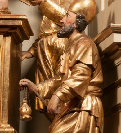 Zdjęcie nr 73: Tabernakulum w formie Arki Przymierza, adorowanej przez dwie klęczące figury starotestamentowego arcykapłana i biskupa oraz dwie stojące postacie lewitów oraz klęczące aniołów, zwieńczonej glorią promienistą z okiem Opatrzności. Wsparte na ośmiu cylindrycznych nóżkach prostopadłościenne tabernakulum jest od frontu podzielone na trzy pola wyznaczone kanelowanymi listwami. Pole środkowe stanowią drzwiczki z rytymi cyframi Dekalogu z ukrzyżowanym Chrystusem, pola boczne z wiązką pałek wodnych z jednej i winnym gronem z drugiej strony. Na skłębionych chmurach z podestem leży Baranek (obecnie w jego miejscu znajduje się figura Matki Boskiej). W tle znacznej wielkości gloria promienista z wieńcem chmur i umieszczonym w nim okiem Opatrzności oraz dwie uskrzydlone główki, nad którymi papieska tiara ze skrzyżowanymi kluczami. Po bokach Baranka dwa klęczące, uskrzydlone nagie aniołki. Obok tabernakulum dwie klęczące postacie z kadzielnicami w rękach. Postać po lewej w stroju biskupim, w albie, stule i kapie oraz infule z dwiema taśmami. Postać po prawej stronie jest ubrany w tradycyjny starotestamentowy strój – efod czyli tunikę i płaszcz wierzchni przepasany na biodrach, pektorał na piersi i zawój na głowie. Za nimi dwaj stojący lewici w tunikach przepasanych na biodrach i wysokich butach. Adorujące Baranka anioły w pozycji klęczącej z rękami skrzyżowanymi na piersi, ze skrzydłami na plecach i nogami w połowie utworzonymi ze skrzydeł.  


