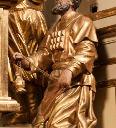 Zdjęcie nr 75: Tabernakulum w formie Arki Przymierza, adorowanej przez dwie klęczące figury starotestamentowego arcykapłana i biskupa oraz dwie stojące postacie lewitów oraz klęczące aniołów, zwieńczonej glorią promienistą z okiem Opatrzności. Wsparte na ośmiu cylindrycznych nóżkach prostopadłościenne tabernakulum jest od frontu podzielone na trzy pola wyznaczone kanelowanymi listwami. Pole środkowe stanowią drzwiczki z rytymi cyframi Dekalogu z ukrzyżowanym Chrystusem, pola boczne z wiązką pałek wodnych z jednej i winnym gronem z drugiej strony. Na skłębionych chmurach z podestem leży Baranek (obecnie w jego miejscu znajduje się figura Matki Boskiej). W tle znacznej wielkości gloria promienista z wieńcem chmur i umieszczonym w nim okiem Opatrzności oraz dwie uskrzydlone główki, nad którymi papieska tiara ze skrzyżowanymi kluczami. Po bokach Baranka dwa klęczące, uskrzydlone nagie aniołki. Obok tabernakulum dwie klęczące postacie z kadzielnicami w rękach. Postać po lewej w stroju biskupim, w albie, stule i kapie oraz infule z dwiema taśmami. Postać po prawej stronie jest ubrany w tradycyjny starotestamentowy strój – efod czyli tunikę i płaszcz wierzchni przepasany na biodrach, pektorał na piersi i zawój na głowie. Za nimi dwaj stojący lewici w tunikach przepasanych na biodrach i wysokich butach. Adorujące Baranka anioły w pozycji klęczącej z rękami skrzyżowanymi na piersi, ze skrzydłami na plecach i nogami w połowie utworzonymi ze skrzydeł.  


