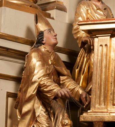 Zdjęcie nr 77: Tabernakulum w formie Arki Przymierza, adorowanej przez dwie klęczące figury starotestamentowego arcykapłana i biskupa oraz dwie stojące postacie lewitów oraz klęczące aniołów, zwieńczonej glorią promienistą z okiem Opatrzności. Wsparte na ośmiu cylindrycznych nóżkach prostopadłościenne tabernakulum jest od frontu podzielone na trzy pola wyznaczone kanelowanymi listwami. Pole środkowe stanowią drzwiczki z rytymi cyframi Dekalogu z ukrzyżowanym Chrystusem, pola boczne z wiązką pałek wodnych z jednej i winnym gronem z drugiej strony. Na skłębionych chmurach z podestem leży Baranek (obecnie w jego miejscu znajduje się figura Matki Boskiej). W tle znacznej wielkości gloria promienista z wieńcem chmur i umieszczonym w nim okiem Opatrzności oraz dwie uskrzydlone główki, nad którymi papieska tiara ze skrzyżowanymi kluczami. Po bokach Baranka dwa klęczące, uskrzydlone nagie aniołki. Obok tabernakulum dwie klęczące postacie z kadzielnicami w rękach. Postać po lewej w stroju biskupim, w albie, stule i kapie oraz infule z dwiema taśmami. Postać po prawej stronie jest ubrany w tradycyjny starotestamentowy strój – efod czyli tunikę i płaszcz wierzchni przepasany na biodrach, pektorał na piersi i zawój na głowie. Za nimi dwaj stojący lewici w tunikach przepasanych na biodrach i wysokich butach. Adorujące Baranka anioły w pozycji klęczącej z rękami skrzyżowanymi na piersi, ze skrzydłami na plecach i nogami w połowie utworzonymi ze skrzydeł.  


