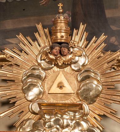 Zdjęcie nr 74: Tabernakulum w formie Arki Przymierza, adorowanej przez dwie klęczące figury starotestamentowego arcykapłana i biskupa oraz dwie stojące postacie lewitów oraz klęczące aniołów, zwieńczonej glorią promienistą z okiem Opatrzności. Wsparte na ośmiu cylindrycznych nóżkach prostopadłościenne tabernakulum jest od frontu podzielone na trzy pola wyznaczone kanelowanymi listwami. Pole środkowe stanowią drzwiczki z rytymi cyframi Dekalogu z ukrzyżowanym Chrystusem, pola boczne z wiązką pałek wodnych z jednej i winnym gronem z drugiej strony. Na skłębionych chmurach z podestem leży Baranek (obecnie w jego miejscu znajduje się figura Matki Boskiej). W tle znacznej wielkości gloria promienista z wieńcem chmur i umieszczonym w nim okiem Opatrzności oraz dwie uskrzydlone główki, nad którymi papieska tiara ze skrzyżowanymi kluczami. Po bokach Baranka dwa klęczące, uskrzydlone nagie aniołki. Obok tabernakulum dwie klęczące postacie z kadzielnicami w rękach. Postać po lewej w stroju biskupim, w albie, stule i kapie oraz infule z dwiema taśmami. Postać po prawej stronie jest ubrany w tradycyjny starotestamentowy strój – efod czyli tunikę i płaszcz wierzchni przepasany na biodrach, pektorał na piersi i zawój na głowie. Za nimi dwaj stojący lewici w tunikach przepasanych na biodrach i wysokich butach. Adorujące Baranka anioły w pozycji klęczącej z rękami skrzyżowanymi na piersi, ze skrzydłami na plecach i nogami w połowie utworzonymi ze skrzydeł.  


