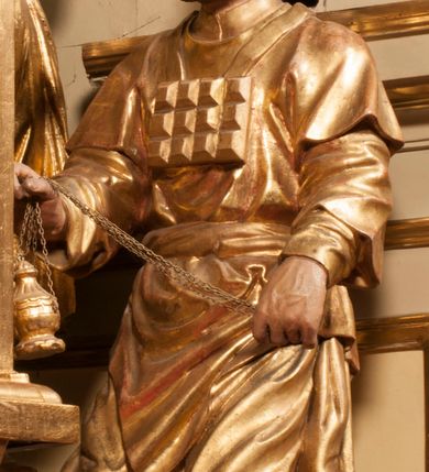 Zdjęcie nr 76: Tabernakulum w formie Arki Przymierza, adorowanej przez dwie klęczące figury starotestamentowego arcykapłana i biskupa oraz dwie stojące postacie lewitów oraz klęczące aniołów, zwieńczonej glorią promienistą z okiem Opatrzności. Wsparte na ośmiu cylindrycznych nóżkach prostopadłościenne tabernakulum jest od frontu podzielone na trzy pola wyznaczone kanelowanymi listwami. Pole środkowe stanowią drzwiczki z rytymi cyframi Dekalogu z ukrzyżowanym Chrystusem, pola boczne z wiązką pałek wodnych z jednej i winnym gronem z drugiej strony. Na skłębionych chmurach z podestem leży Baranek (obecnie w jego miejscu znajduje się figura Matki Boskiej). W tle znacznej wielkości gloria promienista z wieńcem chmur i umieszczonym w nim okiem Opatrzności oraz dwie uskrzydlone główki, nad którymi papieska tiara ze skrzyżowanymi kluczami. Po bokach Baranka dwa klęczące, uskrzydlone nagie aniołki. Obok tabernakulum dwie klęczące postacie z kadzielnicami w rękach. Postać po lewej w stroju biskupim, w albie, stule i kapie oraz infule z dwiema taśmami. Postać po prawej stronie jest ubrany w tradycyjny starotestamentowy strój – efod czyli tunikę i płaszcz wierzchni przepasany na biodrach, pektorał na piersi i zawój na głowie. Za nimi dwaj stojący lewici w tunikach przepasanych na biodrach i wysokich butach. Adorujące Baranka anioły w pozycji klęczącej z rękami skrzyżowanymi na piersi, ze skrzydłami na plecach i nogami w połowie utworzonymi ze skrzydeł.  


