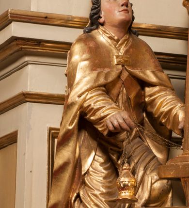 Zdjęcie nr 78: Tabernakulum w formie Arki Przymierza, adorowanej przez dwie klęczące figury starotestamentowego arcykapłana i biskupa oraz dwie stojące postacie lewitów oraz klęczące aniołów, zwieńczonej glorią promienistą z okiem Opatrzności. Wsparte na ośmiu cylindrycznych nóżkach prostopadłościenne tabernakulum jest od frontu podzielone na trzy pola wyznaczone kanelowanymi listwami. Pole środkowe stanowią drzwiczki z rytymi cyframi Dekalogu z ukrzyżowanym Chrystusem, pola boczne z wiązką pałek wodnych z jednej i winnym gronem z drugiej strony. Na skłębionych chmurach z podestem leży Baranek (obecnie w jego miejscu znajduje się figura Matki Boskiej). W tle znacznej wielkości gloria promienista z wieńcem chmur i umieszczonym w nim okiem Opatrzności oraz dwie uskrzydlone główki, nad którymi papieska tiara ze skrzyżowanymi kluczami. Po bokach Baranka dwa klęczące, uskrzydlone nagie aniołki. Obok tabernakulum dwie klęczące postacie z kadzielnicami w rękach. Postać po lewej w stroju biskupim, w albie, stule i kapie oraz infule z dwiema taśmami. Postać po prawej stronie jest ubrany w tradycyjny starotestamentowy strój – efod czyli tunikę i płaszcz wierzchni przepasany na biodrach, pektorał na piersi i zawój na głowie. Za nimi dwaj stojący lewici w tunikach przepasanych na biodrach i wysokich butach. Adorujące Baranka anioły w pozycji klęczącej z rękami skrzyżowanymi na piersi, ze skrzydłami na plecach i nogami w połowie utworzonymi ze skrzydeł.  


