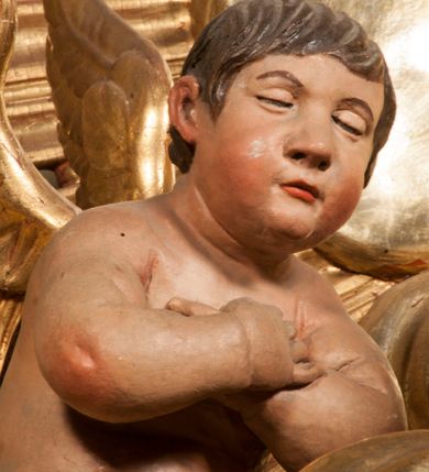 Zdjęcie nr 81: Tabernakulum w formie Arki Przymierza, adorowanej przez dwie klęczące figury starotestamentowego arcykapłana i biskupa oraz dwie stojące postacie lewitów oraz klęczące aniołów, zwieńczonej glorią promienistą z okiem Opatrzności. Wsparte na ośmiu cylindrycznych nóżkach prostopadłościenne tabernakulum jest od frontu podzielone na trzy pola wyznaczone kanelowanymi listwami. Pole środkowe stanowią drzwiczki z rytymi cyframi Dekalogu z ukrzyżowanym Chrystusem, pola boczne z wiązką pałek wodnych z jednej i winnym gronem z drugiej strony. Na skłębionych chmurach z podestem leży Baranek (obecnie w jego miejscu znajduje się figura Matki Boskiej). W tle znacznej wielkości gloria promienista z wieńcem chmur i umieszczonym w nim okiem Opatrzności oraz dwie uskrzydlone główki, nad którymi papieska tiara ze skrzyżowanymi kluczami. Po bokach Baranka dwa klęczące, uskrzydlone nagie aniołki. Obok tabernakulum dwie klęczące postacie z kadzielnicami w rękach. Postać po lewej w stroju biskupim, w albie, stule i kapie oraz infule z dwiema taśmami. Postać po prawej stronie jest ubrany w tradycyjny starotestamentowy strój – efod czyli tunikę i płaszcz wierzchni przepasany na biodrach, pektorał na piersi i zawój na głowie. Za nimi dwaj stojący lewici w tunikach przepasanych na biodrach i wysokich butach. Adorujące Baranka anioły w pozycji klęczącej z rękami skrzyżowanymi na piersi, ze skrzydłami na plecach i nogami w połowie utworzonymi ze skrzydeł.  


