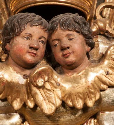 Zdjęcie nr 84: Tabernakulum w formie Arki Przymierza, adorowanej przez dwie klęczące figury starotestamentowego arcykapłana i biskupa oraz dwie stojące postacie lewitów oraz klęczące aniołów, zwieńczonej glorią promienistą z okiem Opatrzności. Wsparte na ośmiu cylindrycznych nóżkach prostopadłościenne tabernakulum jest od frontu podzielone na trzy pola wyznaczone kanelowanymi listwami. Pole środkowe stanowią drzwiczki z rytymi cyframi Dekalogu z ukrzyżowanym Chrystusem, pola boczne z wiązką pałek wodnych z jednej i winnym gronem z drugiej strony. Na skłębionych chmurach z podestem leży Baranek (obecnie w jego miejscu znajduje się figura Matki Boskiej). W tle znacznej wielkości gloria promienista z wieńcem chmur i umieszczonym w nim okiem Opatrzności oraz dwie uskrzydlone główki, nad którymi papieska tiara ze skrzyżowanymi kluczami. Po bokach Baranka dwa klęczące, uskrzydlone nagie aniołki. Obok tabernakulum dwie klęczące postacie z kadzielnicami w rękach. Postać po lewej w stroju biskupim, w albie, stule i kapie oraz infule z dwiema taśmami. Postać po prawej stronie jest ubrany w tradycyjny starotestamentowy strój – efod czyli tunikę i płaszcz wierzchni przepasany na biodrach, pektorał na piersi i zawój na głowie. Za nimi dwaj stojący lewici w tunikach przepasanych na biodrach i wysokich butach. Adorujące Baranka anioły w pozycji klęczącej z rękami skrzyżowanymi na piersi, ze skrzydłami na plecach i nogami w połowie utworzonymi ze skrzydeł.  


