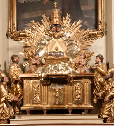 Zdjęcie nr 86: Tabernakulum w formie Arki Przymierza, adorowanej przez dwie klęczące figury starotestamentowego arcykapłana i biskupa oraz dwie stojące postacie lewitów oraz klęczące aniołów, zwieńczonej glorią promienistą z okiem Opatrzności. Wsparte na ośmiu cylindrycznych nóżkach prostopadłościenne tabernakulum jest od frontu podzielone na trzy pola wyznaczone kanelowanymi listwami. Pole środkowe stanowią drzwiczki z rytymi cyframi Dekalogu z ukrzyżowanym Chrystusem, pola boczne z wiązką pałek wodnych z jednej i winnym gronem z drugiej strony. Na skłębionych chmurach z podestem leży Baranek (obecnie w jego miejscu znajduje się figura Matki Boskiej). W tle znacznej wielkości gloria promienista z wieńcem chmur i umieszczonym w nim okiem Opatrzności oraz dwie uskrzydlone główki, nad którymi papieska tiara ze skrzyżowanymi kluczami. Po bokach Baranka dwa klęczące, uskrzydlone nagie aniołki. Obok tabernakulum dwie klęczące postacie z kadzielnicami w rękach. Postać po lewej w stroju biskupim, w albie, stule i kapie oraz infule z dwiema taśmami. Postać po prawej stronie jest ubrany w tradycyjny starotestamentowy strój – efod czyli tunikę i płaszcz wierzchni przepasany na biodrach, pektorał na piersi i zawój na głowie. Za nimi dwaj stojący lewici w tunikach przepasanych na biodrach i wysokich butach. Adorujące Baranka anioły w pozycji klęczącej z rękami skrzyżowanymi na piersi, ze skrzydłami na plecach i nogami w połowie utworzonymi ze skrzydeł.  


