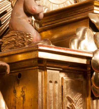 Zdjęcie nr 87: Tabernakulum w formie Arki Przymierza, adorowanej przez dwie klęczące figury starotestamentowego arcykapłana i biskupa oraz dwie stojące postacie lewitów oraz klęczące aniołów, zwieńczonej glorią promienistą z okiem Opatrzności. Wsparte na ośmiu cylindrycznych nóżkach prostopadłościenne tabernakulum jest od frontu podzielone na trzy pola wyznaczone kanelowanymi listwami. Pole środkowe stanowią drzwiczki z rytymi cyframi Dekalogu z ukrzyżowanym Chrystusem, pola boczne z wiązką pałek wodnych z jednej i winnym gronem z drugiej strony. Na skłębionych chmurach z podestem leży Baranek (obecnie w jego miejscu znajduje się figura Matki Boskiej). W tle znacznej wielkości gloria promienista z wieńcem chmur i umieszczonym w nim okiem Opatrzności oraz dwie uskrzydlone główki, nad którymi papieska tiara ze skrzyżowanymi kluczami. Po bokach Baranka dwa klęczące, uskrzydlone nagie aniołki. Obok tabernakulum dwie klęczące postacie z kadzielnicami w rękach. Postać po lewej w stroju biskupim, w albie, stule i kapie oraz infule z dwiema taśmami. Postać po prawej stronie jest ubrany w tradycyjny starotestamentowy strój – efod czyli tunikę i płaszcz wierzchni przepasany na biodrach, pektorał na piersi i zawój na głowie. Za nimi dwaj stojący lewici w tunikach przepasanych na biodrach i wysokich butach. Adorujące Baranka anioły w pozycji klęczącej z rękami skrzyżowanymi na piersi, ze skrzydłami na plecach i nogami w połowie utworzonymi ze skrzydeł.  


