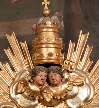 Zdjęcie nr 85: Tabernakulum w formie Arki Przymierza, adorowanej przez dwie klęczące figury starotestamentowego arcykapłana i biskupa oraz dwie stojące postacie lewitów oraz klęczące aniołów, zwieńczonej glorią promienistą z okiem Opatrzności. Wsparte na ośmiu cylindrycznych nóżkach prostopadłościenne tabernakulum jest od frontu podzielone na trzy pola wyznaczone kanelowanymi listwami. Pole środkowe stanowią drzwiczki z rytymi cyframi Dekalogu z ukrzyżowanym Chrystusem, pola boczne z wiązką pałek wodnych z jednej i winnym gronem z drugiej strony. Na skłębionych chmurach z podestem leży Baranek (obecnie w jego miejscu znajduje się figura Matki Boskiej). W tle znacznej wielkości gloria promienista z wieńcem chmur i umieszczonym w nim okiem Opatrzności oraz dwie uskrzydlone główki, nad którymi papieska tiara ze skrzyżowanymi kluczami. Po bokach Baranka dwa klęczące, uskrzydlone nagie aniołki. Obok tabernakulum dwie klęczące postacie z kadzielnicami w rękach. Postać po lewej w stroju biskupim, w albie, stule i kapie oraz infule z dwiema taśmami. Postać po prawej stronie jest ubrany w tradycyjny starotestamentowy strój – efod czyli tunikę i płaszcz wierzchni przepasany na biodrach, pektorał na piersi i zawój na głowie. Za nimi dwaj stojący lewici w tunikach przepasanych na biodrach i wysokich butach. Adorujące Baranka anioły w pozycji klęczącej z rękami skrzyżowanymi na piersi, ze skrzydłami na plecach i nogami w połowie utworzonymi ze skrzydeł.  


