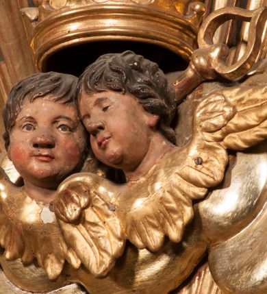 Zdjęcie nr 83: Tabernakulum w formie Arki Przymierza, adorowanej przez dwie klęczące figury starotestamentowego arcykapłana i biskupa oraz dwie stojące postacie lewitów oraz klęczące aniołów, zwieńczonej glorią promienistą z okiem Opatrzności. Wsparte na ośmiu cylindrycznych nóżkach prostopadłościenne tabernakulum jest od frontu podzielone na trzy pola wyznaczone kanelowanymi listwami. Pole środkowe stanowią drzwiczki z rytymi cyframi Dekalogu z ukrzyżowanym Chrystusem, pola boczne z wiązką pałek wodnych z jednej i winnym gronem z drugiej strony. Na skłębionych chmurach z podestem leży Baranek (obecnie w jego miejscu znajduje się figura Matki Boskiej). W tle znacznej wielkości gloria promienista z wieńcem chmur i umieszczonym w nim okiem Opatrzności oraz dwie uskrzydlone główki, nad którymi papieska tiara ze skrzyżowanymi kluczami. Po bokach Baranka dwa klęczące, uskrzydlone nagie aniołki. Obok tabernakulum dwie klęczące postacie z kadzielnicami w rękach. Postać po lewej w stroju biskupim, w albie, stule i kapie oraz infule z dwiema taśmami. Postać po prawej stronie jest ubrany w tradycyjny starotestamentowy strój – efod czyli tunikę i płaszcz wierzchni przepasany na biodrach, pektorał na piersi i zawój na głowie. Za nimi dwaj stojący lewici w tunikach przepasanych na biodrach i wysokich butach. Adorujące Baranka anioły w pozycji klęczącej z rękami skrzyżowanymi na piersi, ze skrzydłami na plecach i nogami w połowie utworzonymi ze skrzydeł.  


