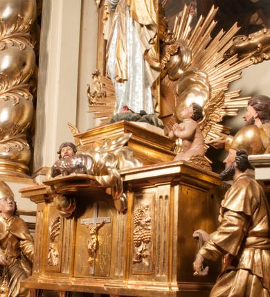 Zdjęcie nr 91: Tabernakulum w formie Arki Przymierza, adorowanej przez dwie klęczące figury starotestamentowego arcykapłana i biskupa oraz dwie stojące postacie lewitów oraz klęczące aniołów, zwieńczonej glorią promienistą z okiem Opatrzności. Wsparte na ośmiu cylindrycznych nóżkach prostopadłościenne tabernakulum jest od frontu podzielone na trzy pola wyznaczone kanelowanymi listwami. Pole środkowe stanowią drzwiczki z rytymi cyframi Dekalogu z ukrzyżowanym Chrystusem, pola boczne z wiązką pałek wodnych z jednej i winnym gronem z drugiej strony. Na skłębionych chmurach z podestem leży Baranek (obecnie w jego miejscu znajduje się figura Matki Boskiej). W tle znacznej wielkości gloria promienista z wieńcem chmur i umieszczonym w nim okiem Opatrzności oraz dwie uskrzydlone główki, nad którymi papieska tiara ze skrzyżowanymi kluczami. Po bokach Baranka dwa klęczące, uskrzydlone nagie aniołki. Obok tabernakulum dwie klęczące postacie z kadzielnicami w rękach. Postać po lewej w stroju biskupim, w albie, stule i kapie oraz infule z dwiema taśmami. Postać po prawej stronie jest ubrany w tradycyjny starotestamentowy strój – efod czyli tunikę i płaszcz wierzchni przepasany na biodrach, pektorał na piersi i zawój na głowie. Za nimi dwaj stojący lewici w tunikach przepasanych na biodrach i wysokich butach. Adorujące Baranka anioły w pozycji klęczącej z rękami skrzyżowanymi na piersi, ze skrzydłami na plecach i nogami w połowie utworzonymi ze skrzydeł.  


