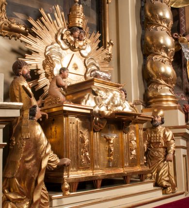 Zdjęcie nr 89: Tabernakulum w formie Arki Przymierza, adorowanej przez dwie klęczące figury starotestamentowego arcykapłana i biskupa oraz dwie stojące postacie lewitów oraz klęczące aniołów, zwieńczonej glorią promienistą z okiem Opatrzności. Wsparte na ośmiu cylindrycznych nóżkach prostopadłościenne tabernakulum jest od frontu podzielone na trzy pola wyznaczone kanelowanymi listwami. Pole środkowe stanowią drzwiczki z rytymi cyframi Dekalogu z ukrzyżowanym Chrystusem, pola boczne z wiązką pałek wodnych z jednej i winnym gronem z drugiej strony. Na skłębionych chmurach z podestem leży Baranek (obecnie w jego miejscu znajduje się figura Matki Boskiej). W tle znacznej wielkości gloria promienista z wieńcem chmur i umieszczonym w nim okiem Opatrzności oraz dwie uskrzydlone główki, nad którymi papieska tiara ze skrzyżowanymi kluczami. Po bokach Baranka dwa klęczące, uskrzydlone nagie aniołki. Obok tabernakulum dwie klęczące postacie z kadzielnicami w rękach. Postać po lewej w stroju biskupim, w albie, stule i kapie oraz infule z dwiema taśmami. Postać po prawej stronie jest ubrany w tradycyjny starotestamentowy strój – efod czyli tunikę i płaszcz wierzchni przepasany na biodrach, pektorał na piersi i zawój na głowie. Za nimi dwaj stojący lewici w tunikach przepasanych na biodrach i wysokich butach. Adorujące Baranka anioły w pozycji klęczącej z rękami skrzyżowanymi na piersi, ze skrzydłami na plecach i nogami w połowie utworzonymi ze skrzydeł.  


