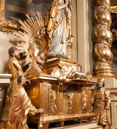 Zdjęcie nr 92: Tabernakulum w formie Arki Przymierza, adorowanej przez dwie klęczące figury starotestamentowego arcykapłana i biskupa oraz dwie stojące postacie lewitów oraz klęczące aniołów, zwieńczonej glorią promienistą z okiem Opatrzności. Wsparte na ośmiu cylindrycznych nóżkach prostopadłościenne tabernakulum jest od frontu podzielone na trzy pola wyznaczone kanelowanymi listwami. Pole środkowe stanowią drzwiczki z rytymi cyframi Dekalogu z ukrzyżowanym Chrystusem, pola boczne z wiązką pałek wodnych z jednej i winnym gronem z drugiej strony. Na skłębionych chmurach z podestem leży Baranek (obecnie w jego miejscu znajduje się figura Matki Boskiej). W tle znacznej wielkości gloria promienista z wieńcem chmur i umieszczonym w nim okiem Opatrzności oraz dwie uskrzydlone główki, nad którymi papieska tiara ze skrzyżowanymi kluczami. Po bokach Baranka dwa klęczące, uskrzydlone nagie aniołki. Obok tabernakulum dwie klęczące postacie z kadzielnicami w rękach. Postać po lewej w stroju biskupim, w albie, stule i kapie oraz infule z dwiema taśmami. Postać po prawej stronie jest ubrany w tradycyjny starotestamentowy strój – efod czyli tunikę i płaszcz wierzchni przepasany na biodrach, pektorał na piersi i zawój na głowie. Za nimi dwaj stojący lewici w tunikach przepasanych na biodrach i wysokich butach. Adorujące Baranka anioły w pozycji klęczącej z rękami skrzyżowanymi na piersi, ze skrzydłami na plecach i nogami w połowie utworzonymi ze skrzydeł.  


