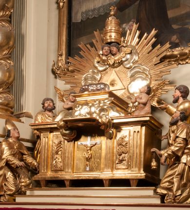 Zdjęcie nr 88: Tabernakulum w formie Arki Przymierza, adorowanej przez dwie klęczące figury starotestamentowego arcykapłana i biskupa oraz dwie stojące postacie lewitów oraz klęczące aniołów, zwieńczonej glorią promienistą z okiem Opatrzności. Wsparte na ośmiu cylindrycznych nóżkach prostopadłościenne tabernakulum jest od frontu podzielone na trzy pola wyznaczone kanelowanymi listwami. Pole środkowe stanowią drzwiczki z rytymi cyframi Dekalogu z ukrzyżowanym Chrystusem, pola boczne z wiązką pałek wodnych z jednej i winnym gronem z drugiej strony. Na skłębionych chmurach z podestem leży Baranek (obecnie w jego miejscu znajduje się figura Matki Boskiej). W tle znacznej wielkości gloria promienista z wieńcem chmur i umieszczonym w nim okiem Opatrzności oraz dwie uskrzydlone główki, nad którymi papieska tiara ze skrzyżowanymi kluczami. Po bokach Baranka dwa klęczące, uskrzydlone nagie aniołki. Obok tabernakulum dwie klęczące postacie z kadzielnicami w rękach. Postać po lewej w stroju biskupim, w albie, stule i kapie oraz infule z dwiema taśmami. Postać po prawej stronie jest ubrany w tradycyjny starotestamentowy strój – efod czyli tunikę i płaszcz wierzchni przepasany na biodrach, pektorał na piersi i zawój na głowie. Za nimi dwaj stojący lewici w tunikach przepasanych na biodrach i wysokich butach. Adorujące Baranka anioły w pozycji klęczącej z rękami skrzyżowanymi na piersi, ze skrzydłami na plecach i nogami w połowie utworzonymi ze skrzydeł.  


