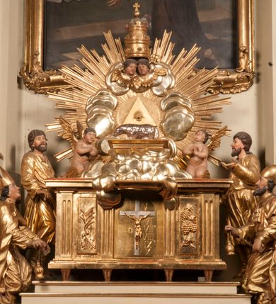 Zdjęcie nr 90: Tabernakulum w formie Arki Przymierza, adorowanej przez dwie klęczące figury starotestamentowego arcykapłana i biskupa oraz dwie stojące postacie lewitów oraz klęczące aniołów, zwieńczonej glorią promienistą z okiem Opatrzności. Wsparte na ośmiu cylindrycznych nóżkach prostopadłościenne tabernakulum jest od frontu podzielone na trzy pola wyznaczone kanelowanymi listwami. Pole środkowe stanowią drzwiczki z rytymi cyframi Dekalogu z ukrzyżowanym Chrystusem, pola boczne z wiązką pałek wodnych z jednej i winnym gronem z drugiej strony. Na skłębionych chmurach z podestem leży Baranek (obecnie w jego miejscu znajduje się figura Matki Boskiej). W tle znacznej wielkości gloria promienista z wieńcem chmur i umieszczonym w nim okiem Opatrzności oraz dwie uskrzydlone główki, nad którymi papieska tiara ze skrzyżowanymi kluczami. Po bokach Baranka dwa klęczące, uskrzydlone nagie aniołki. Obok tabernakulum dwie klęczące postacie z kadzielnicami w rękach. Postać po lewej w stroju biskupim, w albie, stule i kapie oraz infule z dwiema taśmami. Postać po prawej stronie jest ubrany w tradycyjny starotestamentowy strój – efod czyli tunikę i płaszcz wierzchni przepasany na biodrach, pektorał na piersi i zawój na głowie. Za nimi dwaj stojący lewici w tunikach przepasanych na biodrach i wysokich butach. Adorujące Baranka anioły w pozycji klęczącej z rękami skrzyżowanymi na piersi, ze skrzydłami na plecach i nogami w połowie utworzonymi ze skrzydeł.  


