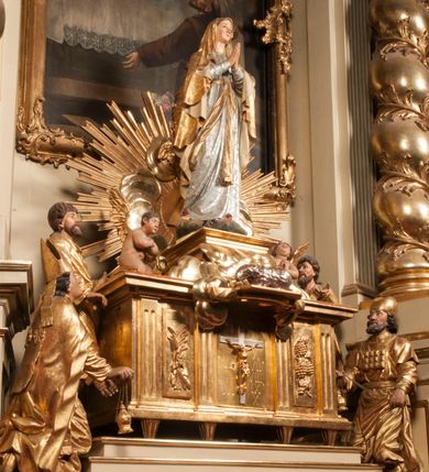 Zdjęcie nr 93: Tabernakulum w formie Arki Przymierza, adorowanej przez dwie klęczące figury starotestamentowego arcykapłana i biskupa oraz dwie stojące postacie lewitów oraz klęczące aniołów, zwieńczonej glorią promienistą z okiem Opatrzności. Wsparte na ośmiu cylindrycznych nóżkach prostopadłościenne tabernakulum jest od frontu podzielone na trzy pola wyznaczone kanelowanymi listwami. Pole środkowe stanowią drzwiczki z rytymi cyframi Dekalogu z ukrzyżowanym Chrystusem, pola boczne z wiązką pałek wodnych z jednej i winnym gronem z drugiej strony. Na skłębionych chmurach z podestem leży Baranek (obecnie w jego miejscu znajduje się figura Matki Boskiej). W tle znacznej wielkości gloria promienista z wieńcem chmur i umieszczonym w nim okiem Opatrzności oraz dwie uskrzydlone główki, nad którymi papieska tiara ze skrzyżowanymi kluczami. Po bokach Baranka dwa klęczące, uskrzydlone nagie aniołki. Obok tabernakulum dwie klęczące postacie z kadzielnicami w rękach. Postać po lewej w stroju biskupim, w albie, stule i kapie oraz infule z dwiema taśmami. Postać po prawej stronie jest ubrany w tradycyjny starotestamentowy strój – efod czyli tunikę i płaszcz wierzchni przepasany na biodrach, pektorał na piersi i zawój na głowie. Za nimi dwaj stojący lewici w tunikach przepasanych na biodrach i wysokich butach. Adorujące Baranka anioły w pozycji klęczącej z rękami skrzyżowanymi na piersi, ze skrzydłami na plecach i nogami w połowie utworzonymi ze skrzydeł.  


