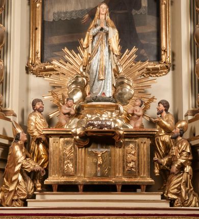 Zdjęcie nr 94: Tabernakulum w formie Arki Przymierza, adorowanej przez dwie klęczące figury starotestamentowego arcykapłana i biskupa oraz dwie stojące postacie lewitów oraz klęczące aniołów, zwieńczonej glorią promienistą z okiem Opatrzności. Wsparte na ośmiu cylindrycznych nóżkach prostopadłościenne tabernakulum jest od frontu podzielone na trzy pola wyznaczone kanelowanymi listwami. Pole środkowe stanowią drzwiczki z rytymi cyframi Dekalogu z ukrzyżowanym Chrystusem, pola boczne z wiązką pałek wodnych z jednej i winnym gronem z drugiej strony. Na skłębionych chmurach z podestem leży Baranek (obecnie w jego miejscu znajduje się figura Matki Boskiej). W tle znacznej wielkości gloria promienista z wieńcem chmur i umieszczonym w nim okiem Opatrzności oraz dwie uskrzydlone główki, nad którymi papieska tiara ze skrzyżowanymi kluczami. Po bokach Baranka dwa klęczące, uskrzydlone nagie aniołki. Obok tabernakulum dwie klęczące postacie z kadzielnicami w rękach. Postać po lewej w stroju biskupim, w albie, stule i kapie oraz infule z dwiema taśmami. Postać po prawej stronie jest ubrany w tradycyjny starotestamentowy strój – efod czyli tunikę i płaszcz wierzchni przepasany na biodrach, pektorał na piersi i zawój na głowie. Za nimi dwaj stojący lewici w tunikach przepasanych na biodrach i wysokich butach. Adorujące Baranka anioły w pozycji klęczącej z rękami skrzyżowanymi na piersi, ze skrzydłami na plecach i nogami w połowie utworzonymi ze skrzydeł.  


