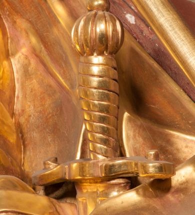 Zdjęcie nr 12: Rzeźba zwrócona w trzech czwartych w prawo, z uniesioną głową, o esowato wygiętej sylwetce, lewą nogę opiera na fragmencie płaszcza. Święty Florian w lewej uniesionej ręce trzyma drzewce rozwianej chorągwi z wizerunkiem orła w koronie, prawą wylewa z dzbana wodę na płonący budynek kościoła stojący przy jego prawej nodze. Twarz ma szczupłą, o dużych, głęboko rzeźbionych oczach, skierowanych w górę i uniesionych, zmarszczonych brwiach, dużym, lekko garbatym i zadartym nosie oraz rozchylonych ustach, okoloną krótką, silnie kręconą brodą oraz rozwianymi na boki włosami. Ubrany jest w krótką tunikę oraz zbroję torsową, przez biodra ma przewiązany pas z zawieszonym przy lewym boku mieczem. Przez lewe ramię ma zawiązany diagonalnie płaszcz, opadający na prawe przedramię oraz zawinięty na lewe udo. Na głowie ma hełm dekorowany maszkaronem z pióropuszem, a na nogach wysokie buty. Polichromia ciała naturalistyczna, włosy brązowe, strój i atrybuty złocone, detale srebrzone. 

