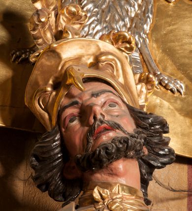 Zdjęcie nr 6: Rzeźba zwrócona w trzech czwartych w prawo, z uniesioną głową, o esowato wygiętej sylwetce, lewą nogę opiera na fragmencie płaszcza. Święty Florian w lewej uniesionej ręce trzyma drzewce rozwianej chorągwi z wizerunkiem orła w koronie, prawą wylewa z dzbana wodę na płonący budynek kościoła stojący przy jego prawej nodze. Twarz ma szczupłą, o dużych, głęboko rzeźbionych oczach, skierowanych w górę i uniesionych, zmarszczonych brwiach, dużym, lekko garbatym i zadartym nosie oraz rozchylonych ustach, okoloną krótką, silnie kręconą brodą oraz rozwianymi na boki włosami. Ubrany jest w krótką tunikę oraz zbroję torsową, przez biodra ma przewiązany pas z zawieszonym przy lewym boku mieczem. Przez lewe ramię ma zawiązany diagonalnie płaszcz, opadający na prawe przedramię oraz zawinięty na lewe udo. Na głowie ma hełm dekorowany maszkaronem z pióropuszem, a na nogach wysokie buty. Polichromia ciała naturalistyczna, włosy brązowe, strój i atrybuty złocone, detale srebrzone. 

