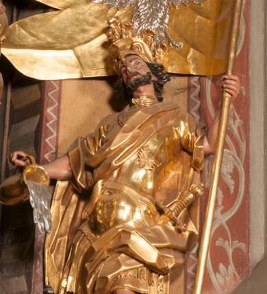 Zdjęcie nr 1: Rzeźba zwrócona w trzech czwartych w prawo, z uniesioną głową, o esowato wygiętej sylwetce, lewą nogę opiera na fragmencie płaszcza. Święty Florian w lewej uniesionej ręce trzyma drzewce rozwianej chorągwi z wizerunkiem orła w koronie, prawą wylewa z dzbana wodę na płonący budynek kościoła stojący przy jego prawej nodze. Twarz ma szczupłą, o dużych, głęboko rzeźbionych oczach, skierowanych w górę i uniesionych, zmarszczonych brwiach, dużym, lekko garbatym i zadartym nosie oraz rozchylonych ustach, okoloną krótką, silnie kręconą brodą oraz rozwianymi na boki włosami. Ubrany jest w krótką tunikę oraz zbroję torsową, przez biodra ma przewiązany pas z zawieszonym przy lewym boku mieczem. Przez lewe ramię ma zawiązany diagonalnie płaszcz, opadający na prawe przedramię oraz zawinięty na lewe udo. Na głowie ma hełm dekorowany maszkaronem z pióropuszem, a na nogach wysokie buty. Polichromia ciała naturalistyczna, włosy brązowe, strój i atrybuty złocone, detale srebrzone. 

