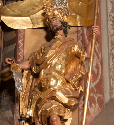 Zdjęcie nr 3: Rzeźba zwrócona w trzech czwartych w prawo, z uniesioną głową, o esowato wygiętej sylwetce, lewą nogę opiera na fragmencie płaszcza. Święty Florian w lewej uniesionej ręce trzyma drzewce rozwianej chorągwi z wizerunkiem orła w koronie, prawą wylewa z dzbana wodę na płonący budynek kościoła stojący przy jego prawej nodze. Twarz ma szczupłą, o dużych, głęboko rzeźbionych oczach, skierowanych w górę i uniesionych, zmarszczonych brwiach, dużym, lekko garbatym i zadartym nosie oraz rozchylonych ustach, okoloną krótką, silnie kręconą brodą oraz rozwianymi na boki włosami. Ubrany jest w krótką tunikę oraz zbroję torsową, przez biodra ma przewiązany pas z zawieszonym przy lewym boku mieczem. Przez lewe ramię ma zawiązany diagonalnie płaszcz, opadający na prawe przedramię oraz zawinięty na lewe udo. Na głowie ma hełm dekorowany maszkaronem z pióropuszem, a na nogach wysokie buty. Polichromia ciała naturalistyczna, włosy brązowe, strój i atrybuty złocone, detale srebrzone. 

