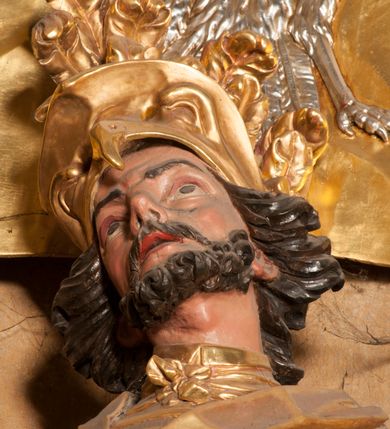 Zdjęcie nr 5: Rzeźba zwrócona w trzech czwartych w prawo, z uniesioną głową, o esowato wygiętej sylwetce, lewą nogę opiera na fragmencie płaszcza. Święty Florian w lewej uniesionej ręce trzyma drzewce rozwianej chorągwi z wizerunkiem orła w koronie, prawą wylewa z dzbana wodę na płonący budynek kościoła stojący przy jego prawej nodze. Twarz ma szczupłą, o dużych, głęboko rzeźbionych oczach, skierowanych w górę i uniesionych, zmarszczonych brwiach, dużym, lekko garbatym i zadartym nosie oraz rozchylonych ustach, okoloną krótką, silnie kręconą brodą oraz rozwianymi na boki włosami. Ubrany jest w krótką tunikę oraz zbroję torsową, przez biodra ma przewiązany pas z zawieszonym przy lewym boku mieczem. Przez lewe ramię ma zawiązany diagonalnie płaszcz, opadający na prawe przedramię oraz zawinięty na lewe udo. Na głowie ma hełm dekorowany maszkaronem z pióropuszem, a na nogach wysokie buty. Polichromia ciała naturalistyczna, włosy brązowe, strój i atrybuty złocone, detale srebrzone. 

