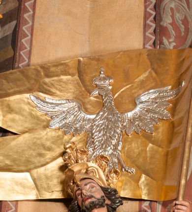 Zdjęcie nr 13: Rzeźba zwrócona w trzech czwartych w prawo, z uniesioną głową, o esowato wygiętej sylwetce, lewą nogę opiera na fragmencie płaszcza. Święty Florian w lewej uniesionej ręce trzyma drzewce rozwianej chorągwi z wizerunkiem orła w koronie, prawą wylewa z dzbana wodę na płonący budynek kościoła stojący przy jego prawej nodze. Twarz ma szczupłą, o dużych, głęboko rzeźbionych oczach, skierowanych w górę i uniesionych, zmarszczonych brwiach, dużym, lekko garbatym i zadartym nosie oraz rozchylonych ustach, okoloną krótką, silnie kręconą brodą oraz rozwianymi na boki włosami. Ubrany jest w krótką tunikę oraz zbroję torsową, przez biodra ma przewiązany pas z zawieszonym przy lewym boku mieczem. Przez lewe ramię ma zawiązany diagonalnie płaszcz, opadający na prawe przedramię oraz zawinięty na lewe udo. Na głowie ma hełm dekorowany maszkaronem z pióropuszem, a na nogach wysokie buty. Polichromia ciała naturalistyczna, włosy brązowe, strój i atrybuty złocone, detale srebrzone. 

