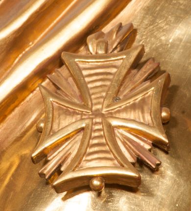 Zdjęcie nr 15: Rzeźba zwrócona w trzech czwartych w prawo, z uniesioną głową, o esowato wygiętej sylwetce, lewą nogę opiera na fragmencie płaszcza. Święty Florian w lewej uniesionej ręce trzyma drzewce rozwianej chorągwi z wizerunkiem orła w koronie, prawą wylewa z dzbana wodę na płonący budynek kościoła stojący przy jego prawej nodze. Twarz ma szczupłą, o dużych, głęboko rzeźbionych oczach, skierowanych w górę i uniesionych, zmarszczonych brwiach, dużym, lekko garbatym i zadartym nosie oraz rozchylonych ustach, okoloną krótką, silnie kręconą brodą oraz rozwianymi na boki włosami. Ubrany jest w krótką tunikę oraz zbroję torsową, przez biodra ma przewiązany pas z zawieszonym przy lewym boku mieczem. Przez lewe ramię ma zawiązany diagonalnie płaszcz, opadający na prawe przedramię oraz zawinięty na lewe udo. Na głowie ma hełm dekorowany maszkaronem z pióropuszem, a na nogach wysokie buty. Polichromia ciała naturalistyczna, włosy brązowe, strój i atrybuty złocone, detale srebrzone. 

