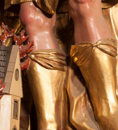 Zdjęcie nr 19: Rzeźba zwrócona w trzech czwartych w prawo, z uniesioną głową, o esowato wygiętej sylwetce, lewą nogę opiera na fragmencie płaszcza. Święty Florian w lewej uniesionej ręce trzyma drzewce rozwianej chorągwi z wizerunkiem orła w koronie, prawą wylewa z dzbana wodę na płonący budynek kościoła stojący przy jego prawej nodze. Twarz ma szczupłą, o dużych, głęboko rzeźbionych oczach, skierowanych w górę i uniesionych, zmarszczonych brwiach, dużym, lekko garbatym i zadartym nosie oraz rozchylonych ustach, okoloną krótką, silnie kręconą brodą oraz rozwianymi na boki włosami. Ubrany jest w krótką tunikę oraz zbroję torsową, przez biodra ma przewiązany pas z zawieszonym przy lewym boku mieczem. Przez lewe ramię ma zawiązany diagonalnie płaszcz, opadający na prawe przedramię oraz zawinięty na lewe udo. Na głowie ma hełm dekorowany maszkaronem z pióropuszem, a na nogach wysokie buty. Polichromia ciała naturalistyczna, włosy brązowe, strój i atrybuty złocone, detale srebrzone. 

