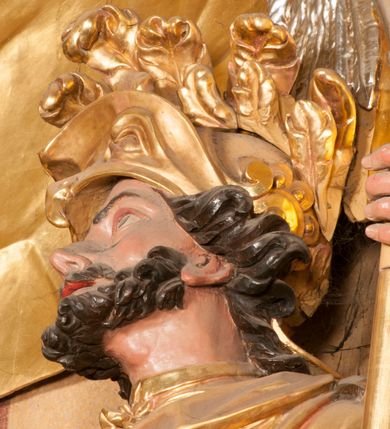 Zdjęcie nr 8: Rzeźba zwrócona w trzech czwartych w prawo, z uniesioną głową, o esowato wygiętej sylwetce, lewą nogę opiera na fragmencie płaszcza. Święty Florian w lewej uniesionej ręce trzyma drzewce rozwianej chorągwi z wizerunkiem orła w koronie, prawą wylewa z dzbana wodę na płonący budynek kościoła stojący przy jego prawej nodze. Twarz ma szczupłą, o dużych, głęboko rzeźbionych oczach, skierowanych w górę i uniesionych, zmarszczonych brwiach, dużym, lekko garbatym i zadartym nosie oraz rozchylonych ustach, okoloną krótką, silnie kręconą brodą oraz rozwianymi na boki włosami. Ubrany jest w krótką tunikę oraz zbroję torsową, przez biodra ma przewiązany pas z zawieszonym przy lewym boku mieczem. Przez lewe ramię ma zawiązany diagonalnie płaszcz, opadający na prawe przedramię oraz zawinięty na lewe udo. Na głowie ma hełm dekorowany maszkaronem z pióropuszem, a na nogach wysokie buty. Polichromia ciała naturalistyczna, włosy brązowe, strój i atrybuty złocone, detale srebrzone. 

