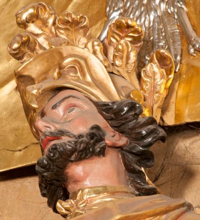 Zdjęcie nr 9: Rzeźba zwrócona w trzech czwartych w prawo, z uniesioną głową, o esowato wygiętej sylwetce, lewą nogę opiera na fragmencie płaszcza. Święty Florian w lewej uniesionej ręce trzyma drzewce rozwianej chorągwi z wizerunkiem orła w koronie, prawą wylewa z dzbana wodę na płonący budynek kościoła stojący przy jego prawej nodze. Twarz ma szczupłą, o dużych, głęboko rzeźbionych oczach, skierowanych w górę i uniesionych, zmarszczonych brwiach, dużym, lekko garbatym i zadartym nosie oraz rozchylonych ustach, okoloną krótką, silnie kręconą brodą oraz rozwianymi na boki włosami. Ubrany jest w krótką tunikę oraz zbroję torsową, przez biodra ma przewiązany pas z zawieszonym przy lewym boku mieczem. Przez lewe ramię ma zawiązany diagonalnie płaszcz, opadający na prawe przedramię oraz zawinięty na lewe udo. Na głowie ma hełm dekorowany maszkaronem z pióropuszem, a na nogach wysokie buty. Polichromia ciała naturalistyczna, włosy brązowe, strój i atrybuty złocone, detale srebrzone. 

