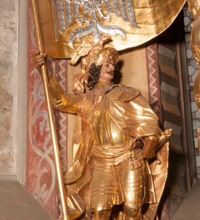 Zdjęcie nr 1: Rzeźba ukazana frontalnie, z głową skierowaną w lewo, w kontrapoście, o esowato wygiętej sylwetce. Lewą ręką trzyma rękojeść miecza przy boku, prawą, uniesioną trzyma drzewce rozwianej chorągwi z wizerunkiem orła (czeska czarna orlica). Twarz ma szczupłą, o dużych oczach, uniesionych brwiach, dużym, zadartym nosie i pełnych ustach, z wąsami, okoloną silnie kręconymi w osobne pukle włosami, sięgającymi ramion. Święty jest ubrany w zbroję płytową, przepasaną w talii szarfą z dwoma chwostami i mieczem przy lewym boku. Na lewym ramieniu ma zawieszony płaszcz opadający z tyłu postaci, a na piersi krzyż kawalerski na taśmie. Na głowie ma hełm z maszkaronem z przodu, zwieńczony pegazem. Polichromia ciała naturalistyczna, włosy brązowe, strój i atrybuty złocone, orzeł srebrzony. 



