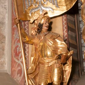 Zdjęcie nr 1: Rzeźba ukazana frontalnie, z głową skierowaną w lewo, w kontrapoście, o esowato wygiętej sylwetce. Lewą ręką trzyma rękojeść miecza przy boku, prawą, uniesioną trzyma drzewce rozwianej chorągwi z wizerunkiem orła (czeska czarna orlica). Twarz ma szczupłą, o dużych oczach, uniesionych brwiach, dużym, zadartym nosie i pełnych ustach, z wąsami, okoloną silnie kręconymi w osobne pukle włosami, sięgającymi ramion. Święty jest ubrany w zbroję płytową, przepasaną w talii szarfą z dwoma chwostami i mieczem przy lewym boku. Na lewym ramieniu ma zawieszony płaszcz opadający z tyłu postaci, a na piersi krzyż kawalerski na taśmie. Na głowie ma hełm z maszkaronem z przodu, zwieńczony pegazem. Polichromia ciała naturalistyczna, włosy brązowe, strój i atrybuty złocone, orzeł srebrzony. 



