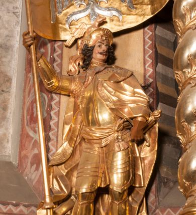 Zdjęcie nr 4: Rzeźba ukazana frontalnie, z głową skierowaną w lewo, w kontrapoście, o esowato wygiętej sylwetce. Lewą ręką trzyma rękojeść miecza przy boku, prawą, uniesioną trzyma drzewce rozwianej chorągwi z wizerunkiem orła (czeska czarna orlica). Twarz ma szczupłą, o dużych oczach, uniesionych brwiach, dużym, zadartym nosie i pełnych ustach, z wąsami, okoloną silnie kręconymi w osobne pukle włosami, sięgającymi ramion. Święty jest ubrany w zbroję płytową, przepasaną w talii szarfą z dwoma chwostami i mieczem przy lewym boku. Na lewym ramieniu ma zawieszony płaszcz opadający z tyłu postaci, a na piersi krzyż kawalerski na taśmie. Na głowie ma hełm z maszkaronem z przodu, zwieńczony pegazem. Polichromia ciała naturalistyczna, włosy brązowe, strój i atrybuty złocone, orzeł srebrzony. 



