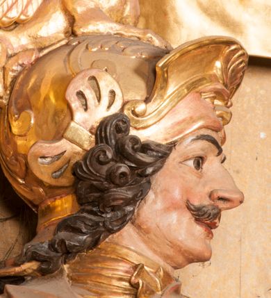 Zdjęcie nr 6: Rzeźba ukazana frontalnie, z głową skierowaną w lewo, w kontrapoście, o esowato wygiętej sylwetce. Lewą ręką trzyma rękojeść miecza przy boku, prawą, uniesioną trzyma drzewce rozwianej chorągwi z wizerunkiem orła (czeska czarna orlica). Twarz ma szczupłą, o dużych oczach, uniesionych brwiach, dużym, zadartym nosie i pełnych ustach, z wąsami, okoloną silnie kręconymi w osobne pukle włosami, sięgającymi ramion. Święty jest ubrany w zbroję płytową, przepasaną w talii szarfą z dwoma chwostami i mieczem przy lewym boku. Na lewym ramieniu ma zawieszony płaszcz opadający z tyłu postaci, a na piersi krzyż kawalerski na taśmie. Na głowie ma hełm z maszkaronem z przodu, zwieńczony pegazem. Polichromia ciała naturalistyczna, włosy brązowe, strój i atrybuty złocone, orzeł srebrzony. 



