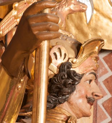 Zdjęcie nr 9: Rzeźba ukazana frontalnie, z głową skierowaną w lewo, w kontrapoście, o esowato wygiętej sylwetce. Lewą ręką trzyma rękojeść miecza przy boku, prawą, uniesioną trzyma drzewce rozwianej chorągwi z wizerunkiem orła (czeska czarna orlica). Twarz ma szczupłą, o dużych oczach, uniesionych brwiach, dużym, zadartym nosie i pełnych ustach, z wąsami, okoloną silnie kręconymi w osobne pukle włosami, sięgającymi ramion. Święty jest ubrany w zbroję płytową, przepasaną w talii szarfą z dwoma chwostami i mieczem przy lewym boku. Na lewym ramieniu ma zawieszony płaszcz opadający z tyłu postaci, a na piersi krzyż kawalerski na taśmie. Na głowie ma hełm z maszkaronem z przodu, zwieńczony pegazem. Polichromia ciała naturalistyczna, włosy brązowe, strój i atrybuty złocone, orzeł srebrzony. 



