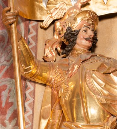 Zdjęcie nr 5: Rzeźba ukazana frontalnie, z głową skierowaną w lewo, w kontrapoście, o esowato wygiętej sylwetce. Lewą ręką trzyma rękojeść miecza przy boku, prawą, uniesioną trzyma drzewce rozwianej chorągwi z wizerunkiem orła (czeska czarna orlica). Twarz ma szczupłą, o dużych oczach, uniesionych brwiach, dużym, zadartym nosie i pełnych ustach, z wąsami, okoloną silnie kręconymi w osobne pukle włosami, sięgającymi ramion. Święty jest ubrany w zbroję płytową, przepasaną w talii szarfą z dwoma chwostami i mieczem przy lewym boku. Na lewym ramieniu ma zawieszony płaszcz opadający z tyłu postaci, a na piersi krzyż kawalerski na taśmie. Na głowie ma hełm z maszkaronem z przodu, zwieńczony pegazem. Polichromia ciała naturalistyczna, włosy brązowe, strój i atrybuty złocone, orzeł srebrzony. 



