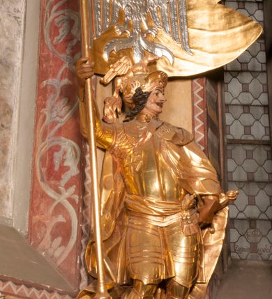 Zdjęcie nr 3: Rzeźba ukazana frontalnie, z głową skierowaną w lewo, w kontrapoście, o esowato wygiętej sylwetce. Lewą ręką trzyma rękojeść miecza przy boku, prawą, uniesioną trzyma drzewce rozwianej chorągwi z wizerunkiem orła (czeska czarna orlica). Twarz ma szczupłą, o dużych oczach, uniesionych brwiach, dużym, zadartym nosie i pełnych ustach, z wąsami, okoloną silnie kręconymi w osobne pukle włosami, sięgającymi ramion. Święty jest ubrany w zbroję płytową, przepasaną w talii szarfą z dwoma chwostami i mieczem przy lewym boku. Na lewym ramieniu ma zawieszony płaszcz opadający z tyłu postaci, a na piersi krzyż kawalerski na taśmie. Na głowie ma hełm z maszkaronem z przodu, zwieńczony pegazem. Polichromia ciała naturalistyczna, włosy brązowe, strój i atrybuty złocone, orzeł srebrzony. 



