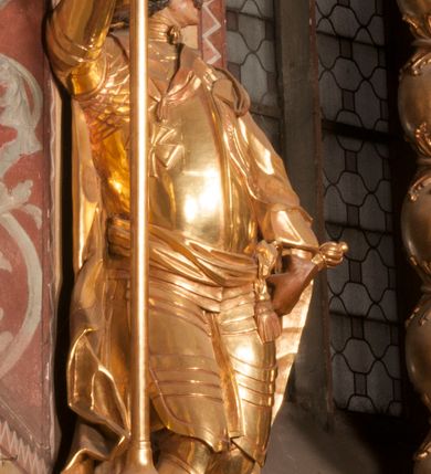 Zdjęcie nr 2: Rzeźba ukazana frontalnie, z głową skierowaną w lewo, w kontrapoście, o esowato wygiętej sylwetce. Lewą ręką trzyma rękojeść miecza przy boku, prawą, uniesioną trzyma drzewce rozwianej chorągwi z wizerunkiem orła (czeska czarna orlica). Twarz ma szczupłą, o dużych oczach, uniesionych brwiach, dużym, zadartym nosie i pełnych ustach, z wąsami, okoloną silnie kręconymi w osobne pukle włosami, sięgającymi ramion. Święty jest ubrany w zbroję płytową, przepasaną w talii szarfą z dwoma chwostami i mieczem przy lewym boku. Na lewym ramieniu ma zawieszony płaszcz opadający z tyłu postaci, a na piersi krzyż kawalerski na taśmie. Na głowie ma hełm z maszkaronem z przodu, zwieńczony pegazem. Polichromia ciała naturalistyczna, włosy brązowe, strój i atrybuty złocone, orzeł srebrzony. 



