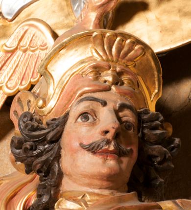 Zdjęcie nr 7: Rzeźba ukazana frontalnie, z głową skierowaną w lewo, w kontrapoście, o esowato wygiętej sylwetce. Lewą ręką trzyma rękojeść miecza przy boku, prawą, uniesioną trzyma drzewce rozwianej chorągwi z wizerunkiem orła (czeska czarna orlica). Twarz ma szczupłą, o dużych oczach, uniesionych brwiach, dużym, zadartym nosie i pełnych ustach, z wąsami, okoloną silnie kręconymi w osobne pukle włosami, sięgającymi ramion. Święty jest ubrany w zbroję płytową, przepasaną w talii szarfą z dwoma chwostami i mieczem przy lewym boku. Na lewym ramieniu ma zawieszony płaszcz opadający z tyłu postaci, a na piersi krzyż kawalerski na taśmie. Na głowie ma hełm z maszkaronem z przodu, zwieńczony pegazem. Polichromia ciała naturalistyczna, włosy brązowe, strój i atrybuty złocone, orzeł srebrzony. 



