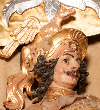 Zdjęcie nr 8: Rzeźba ukazana frontalnie, z głową skierowaną w lewo, w kontrapoście, o esowato wygiętej sylwetce. Lewą ręką trzyma rękojeść miecza przy boku, prawą, uniesioną trzyma drzewce rozwianej chorągwi z wizerunkiem orła (czeska czarna orlica). Twarz ma szczupłą, o dużych oczach, uniesionych brwiach, dużym, zadartym nosie i pełnych ustach, z wąsami, okoloną silnie kręconymi w osobne pukle włosami, sięgającymi ramion. Święty jest ubrany w zbroję płytową, przepasaną w talii szarfą z dwoma chwostami i mieczem przy lewym boku. Na lewym ramieniu ma zawieszony płaszcz opadający z tyłu postaci, a na piersi krzyż kawalerski na taśmie. Na głowie ma hełm z maszkaronem z przodu, zwieńczony pegazem. Polichromia ciała naturalistyczna, włosy brązowe, strój i atrybuty złocone, orzeł srebrzony. 



